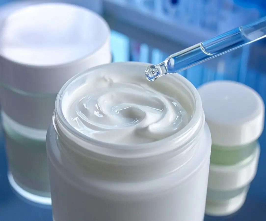 Open cosmetic jar filled with white cream, with other closed jars nearby; a pipette dispensing liquid onto the cream.